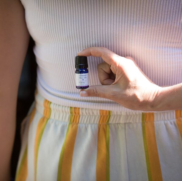 A person holding a small bottle of Kidney Support™ - 5 ml essential oil against their waist, with the label facing outward, to showcase the product.